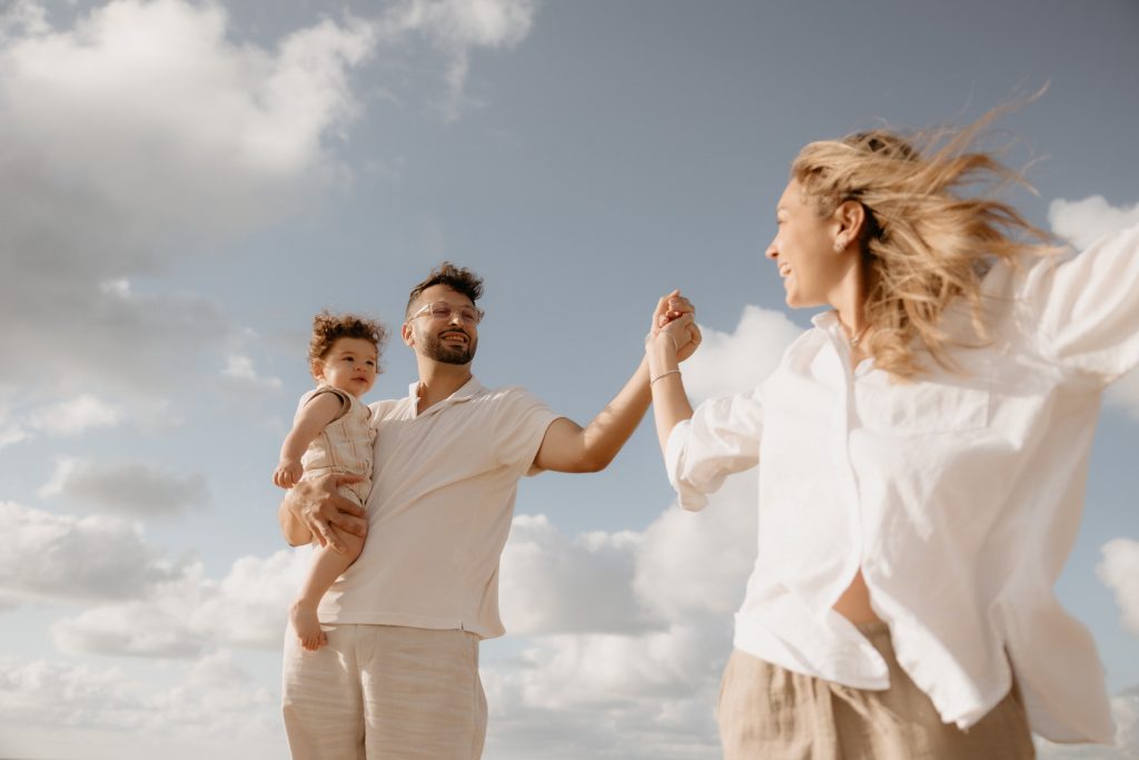 Un couple de parent se tiennent la main et avancent en se regardant. Le papa porte son enfant dans les bras. On voit le beau ciel bleu. Un moment de connexion pris pendant leur séance photo famille par Lesley Sanchez, photographe Landes.