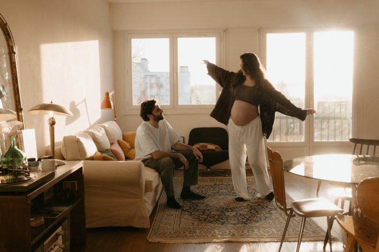Séance grossesse en intérieur. L'homme est assis sur le canapé pendant que la femme enceinte danse au milieu du salon. Un moment fun capturé par Lesley Sanchez, photographe Landes.